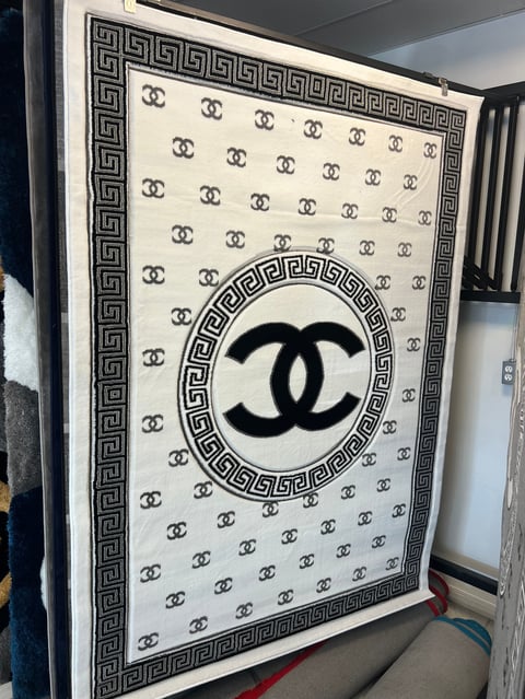 Luxury designer blanket with cream background, repeating interlocking CC logo pattern, black geometric Greek key border, and large centered black CC double-C logo