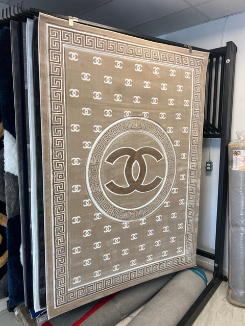 Beige and cream designer rug with CC logo pattern, Greek key border, displayed on display rack